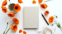 Linen hardcover notebook surrounded by vibrant orange poppy flowers, ink pen, and twine on a white background, flat lay style.