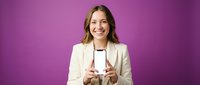 Happy young Caucasian businesswoman in beige blazer holding smartphone with blank white screen on purple background for mobile app mockup presentation.