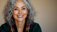 Confident mature woman with natural gray curly hair wearing emerald green sweater and ethnic jewelry smiling warmly at camera against neutral background.