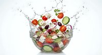Fresh vegetable salad with tomatoes, cucumbers and olives splashing in glass bowl on white background for healthy eating and cooking concepts.