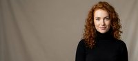 Professional redhead woman with curly hair in black turtleneck sweater smiling confidently against neutral background for business and lifestyle content.