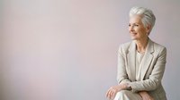 Senior woman with gray hair wearing beige blazer and white shirt, looking to side against neutral background. Corporate portrait for business, leadership, and professional themes.