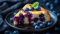 Slice of moist blueberry cake garnished with fresh berries and mint leaves on dark plate. Purple fruit sauce drizzle creates elegant contrast against golden crumb texture.