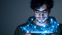 Young Hispanic man with glasses illuminated by blue digital network hologram effect emerging from mobile device, representing future technology and connectivity.