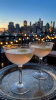 Two elegant cocktail glasses filled with champagne on rooftop terrace overlooking city skyline at sunset with bokeh lights creating romantic atmosphere.