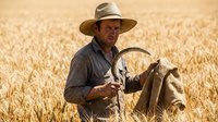 Weathered Caucasian male farmer holding a sickle and burlap sack in a golden wheat field during harvest season under bright summer sun.