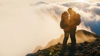 Silhouette of hiking couple embracing on mountain peak at golden sunset with dramatic clouds and lens flare creating romantic outdoor adventure moment.