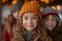 Smiling young girl with curly hair wearing orange knit beanie and winter coat against blurred bokeh lights background, expressing joy and holiday spirit.