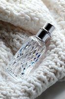 Crystal glass perfume bottle with silver cap on soft white knitted textile background, elegant fragrance container with diamond pattern design and light reflections.