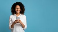 Young African American woman with curly hair smiling while using smartphone on blue background for social media and technology concepts.