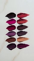 Makeup swatches showing range of dark and bold lipstick colors from deep purple to bright red and nude shades on white marble background for cosmetic advertising.