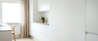 Minimalist white kitchen interior with modern cabinetry, built-in shelving, natural light from window, and potted plant. Suitable for home design, interior decoration, and lifestyle publications.