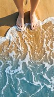 Bare feet standing on golden sandy beach with turquoise ocean waves washing ashore, aerial view of tropical coastline for summer vacation concepts.