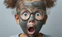 Young African American child wearing round glasses with facial clay mask showing surprised expression with wide open mouth and eyes against gray background.