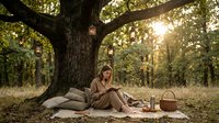 Relaxed woman sitting under large tree in forest clearing with picnic basket and blanket, enjoying peaceful outdoor leisure time surrounded by natural woodland scenery.