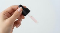 Hand holding glass dropper bottle with black cap dispensing clear liquid drop against white background for skincare cosmetic product mockup.