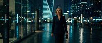 Professional businesswoman walking through modern glass corridor at night with city skyline backdrop. Corporate executive in elegant attire for business concepts.