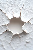 Cracked white wall with hole revealing gray background, showing detailed texture of broken plaster and jagged edges. Dramatic lighting emphasizes depth and damage.