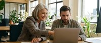 Senior woman with gray hair and a younger man with dark hair and beard, both of Caucasian ethnicity, working together at a laptop in a bright, plant-filled office space. Represents mentorship, collaboration, and intergenerational teamwork in modern workplace.