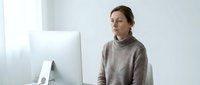 Professional Caucasian woman in beige turtleneck sweater working at computer in modern minimalist office workspace with natural lighting.
