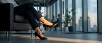 Professional businesswoman in elegant black heels relaxing in modern office lobby during break time. Corporate lifestyle and workplace wellness concept.