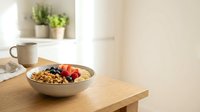 Granola bowl with fresh berries and banana on wooden table in bright kitchen with potted plants and coffee mug nearby.