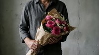 Man in casual shirt holding burgundy rose bouquet wrapped in brown paper against concrete wall background for romantic gesture or special occasion.