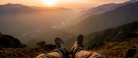 Hiker resting on mountain peak at golden sunset, legs stretched out with hiking boots, panoramic valley and river view in warm light.