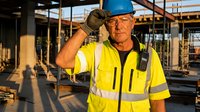 Senior construction worker in safety gear wiping sweat at building site during hot weather conditions. Industrial workplace heat stress and worker fatigue concept.
