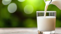 Fresh milk pouring from pitcher into glass on wooden table with green bokeh background, dairy drink photography for healthy lifestyle and nutrition.