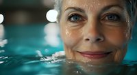Senior woman with bright smile swimming in turquoise pool water, face partially submerged showing vitality and active lifestyle in retirement years.