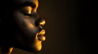 Side profile of African woman with closed eyes in dramatic golden lighting against dark background, highlighting facial features and natural beauty.