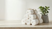 Rolled white towels stacked on wooden surface with green plant in modern concrete pot against neutral wall background for spa wellness bathroom decor.