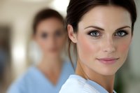 Young female medical professional in white uniform with confident smile and striking blue eyes, with colleague in blue scrubs standing in background at hospital.