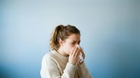 Young woman with light brown hair, Caucasian ethnicity, wearing beige sweater, blowing nose into tissue against blue gradient background. Suitable for healthcare, wellness, cold and flu campaigns.