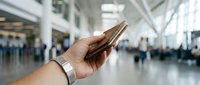 Young adult with medium skin tone holding smartphone in modern airport terminal with blurred travelers and bright natural lighting, suitable for travel, technology, and communication concepts.