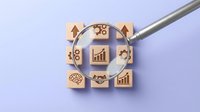 Magnifying glass over wooden blocks with business icons including growth charts, gears, brain, and handshake on light purple background.