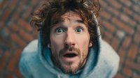 Young Caucasian man with curly hair showing shocked surprised facial expression wearing blue hoodie against brick wall background for advertising campaigns.