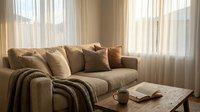 Cozy living room with beige upholstered sofa, decorative pillows, knitted throw blanket, wooden coffee table, and sheer curtains allowing natural light for home decor and interior design projects.