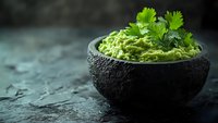 Fresh guacamole dip garnished with cilantro leaves served in black stone bowl on dark textured background with copy space for menu or recipe design.