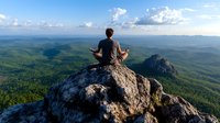 Young man in meditation pose sitting on mountain peak overlooking vast green forest landscape with dramatic clouds and blue sky, practicing mindfulness in nature.