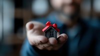 Small gray house model with red bow held gently in palm against blurred dark background, symbolizing home ownership, real estate gift or property investment.