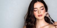 Young woman with closed eyes and natural makeup having blush applied with professional makeup brush against neutral gray background.