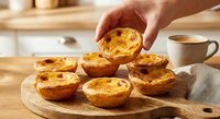 Hand selecting golden baked egg tart from wooden board in warm kitchen setting with coffee cup. Fresh pastries for breakfast or dessert serving.