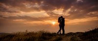 Silhouetted couple embracing on hilltop at golden sunset with dramatic sky and wild grass, romantic outdoor scene for love and relationship content.