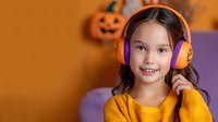 Young girl wearing orange headphones smiling at camera against Halloween background with pumpkin decorations, dressed in bright yellow sweater. Cheerful autumn holiday mood.