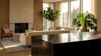 Modern minimalist living room with sleek kitchen island, neutral tones, large windows, and indoor plants creating warm contemporary home interior design.