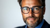 Confident middle-aged Caucasian businessman with gray beard wearing glasses and navy suit jacket smiles warmly against light background in professional headshot portrait.