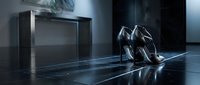 Pair of black high heels displayed on reflective floor next to minimalist dark cabinet in modern interior space, suitable for fashion retail and luxury product marketing.