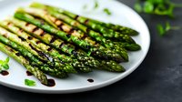 Grilled green asparagus spears on white plate drizzled with balsamic reduction and garnished with fresh herbs against dark background, food photography.